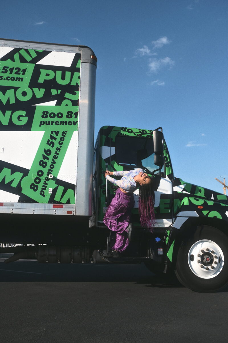 A person with long hair and colorful attire is hanging onto the door of a green and white moving truck with "puremoving.com" branding, against a clear blue sky. For more details or assistance, don't hesitate to Contact Us. Boca Raton Movers