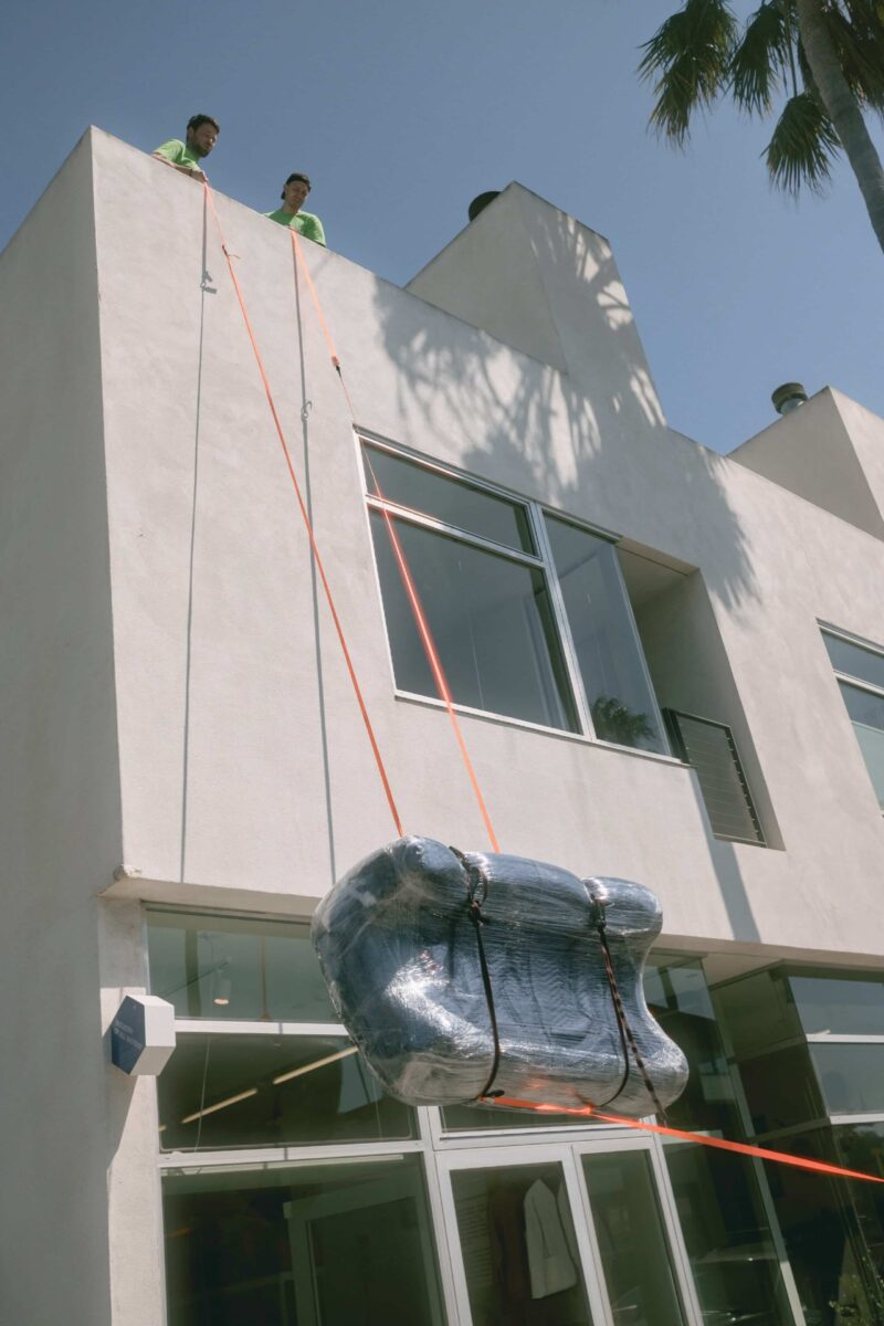 Two people at the top of a building use ropes to lower a large, wrapped object from a window, part of their move from Los Angeles to Miami.