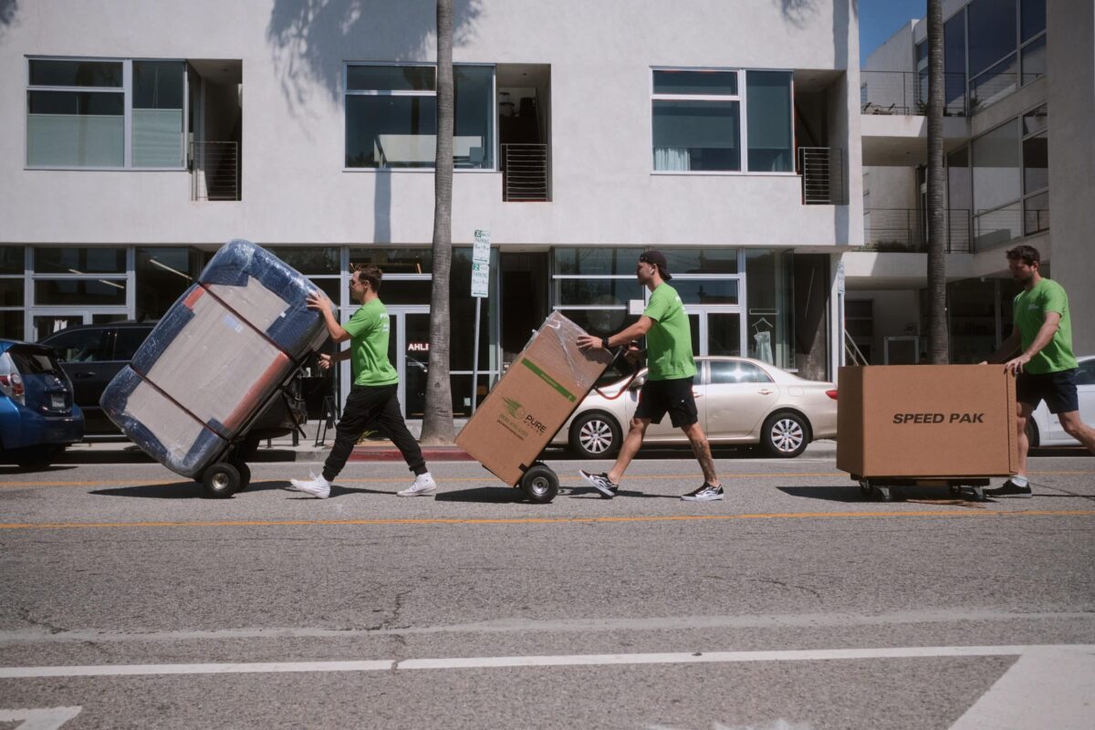 Three movers in green shirts, offering top-notch Moving and Packing Services, push items on dollies across a street in front of a modern apartment building. Pasadena