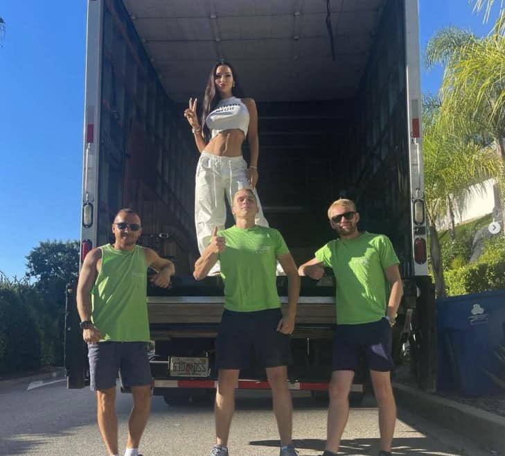 Three men in green shirts and shorts stand in front of a truck, with a woman in white clothing posing inside the truck. The woman, perhaps moving from Los Angeles to Miami, is making a peace sign. Commercial movers