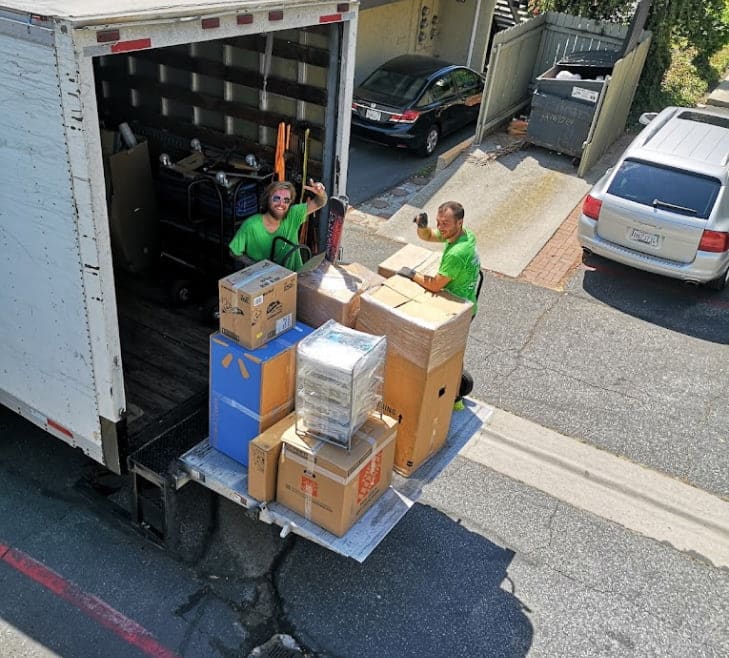 Two movers in green shirts stand on the back of a truck filled with boxes, efficiently organizing items for a Full Service Moving operation on a residential street. Boca Raton Movers