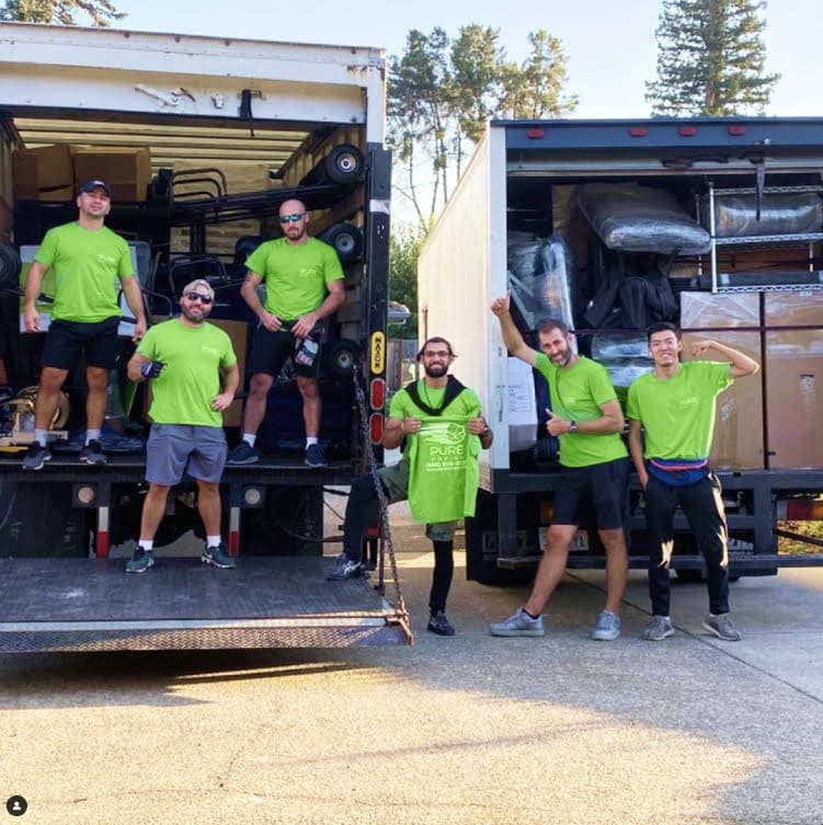 Six men in green shirts stand on and around the back of two moving trucks loaded with items. One man holds a green t-shirt that reads "PURE." With trees visible in the background, this scene encapsulates the efficiency and organization about Bay Area movers.