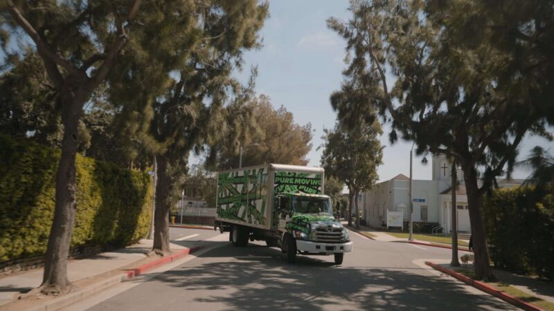 A moving truck painted with green camo patterns and "Pure Moving" text drives through a residential neighborhood lined with tall trees, representing an efficient commercial moving service.