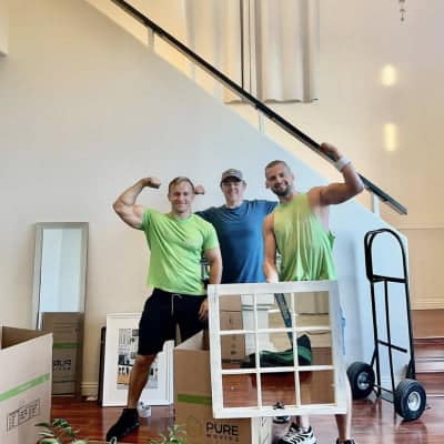 Three men, one in a blue shirt and two in green shirts, pose while flexing their arms in a partially furnished room with moving boxes, a hand truck nearby, and competitive moving rates. Commercial movers