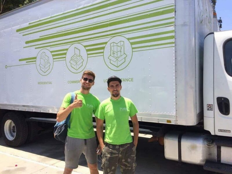 Two movers in green shirts standing in front of a white truck with green graphics, one gesturing a thumbs-up. Long Island