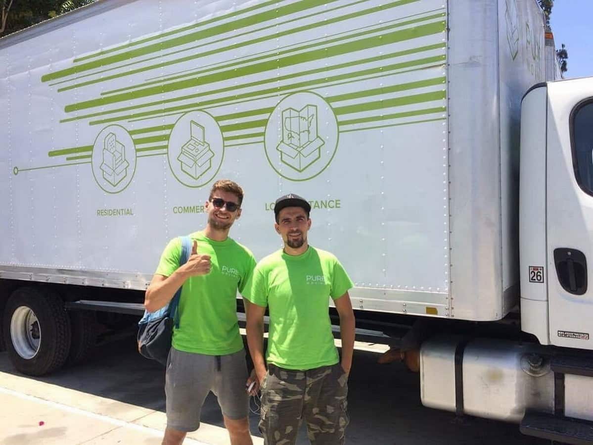 Two movers in green shirts standing in front of a white truck with green graphics, one gesturing a thumbs-up.