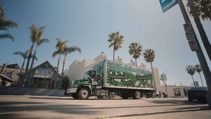 A green and white moving truck is parked on a sunny street lined with palm trees and buildings, signaling someone's exciting journey of moving to Los Angeles. Portlandmovers2