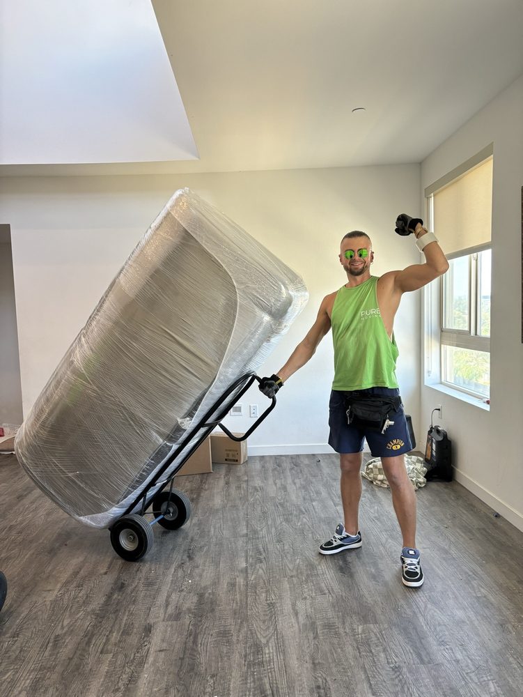 Pure Movers employee transporting wrapped mattress with moving dolly