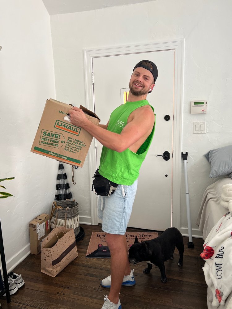 Friendly mover carrying U-Haul moving box inside customer home with dog nearby