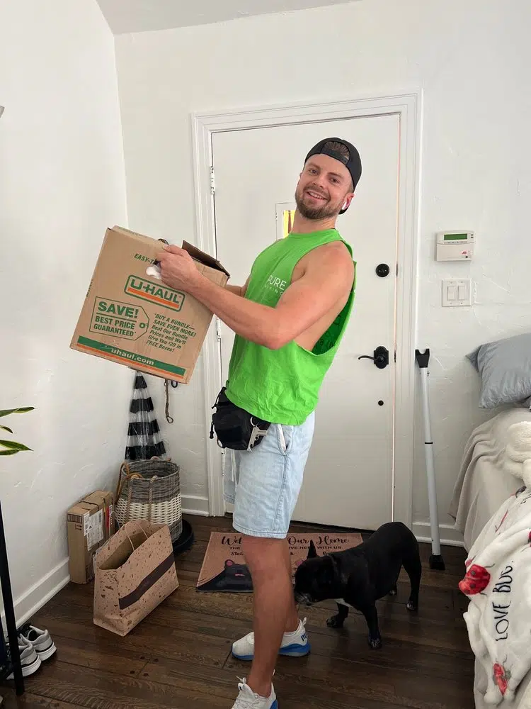 Friendly mover carrying U-Haul moving box inside customer home with dog nearby